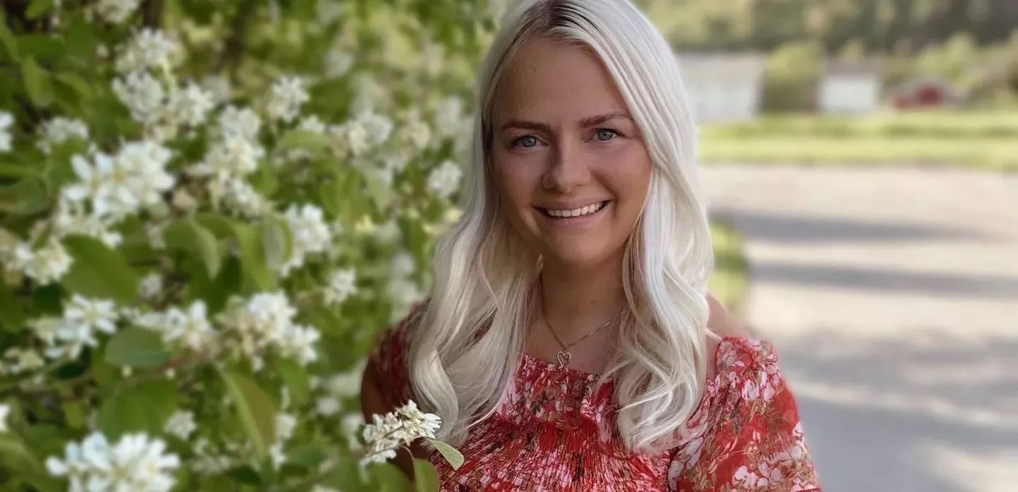 Eine blonde Frau mit sonnengeküsster Haut lächelt vor einem grünen Hintergrund.